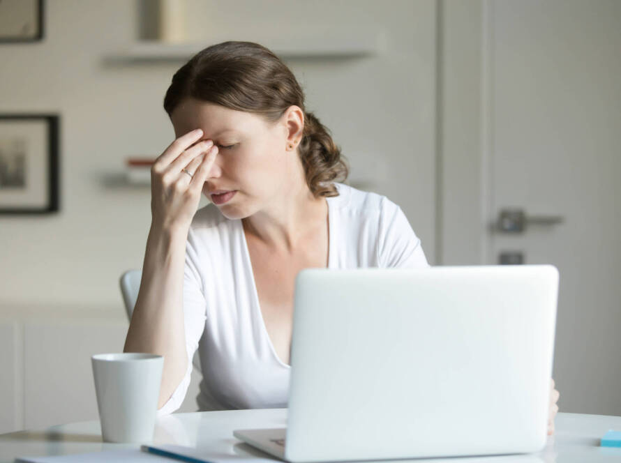 Portrait of a woman on her desk with a laptop, one hand on her forehead.
